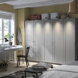 Velký pokoj s vysokými bílými skříněmi PAX. Psací stůl se židlí a okny. Moderní, minimalistický design.