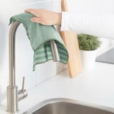 Osoba držící zelený čisticí hadřík pod kuchyňským kohoutkem, s prkénkem a rostlinou v pozadí.