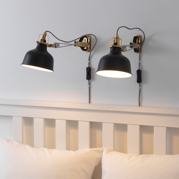 Dvě černé nastavitelné nástěnné lampy RANARP nad bílým dřevěným čelem s polštáři.