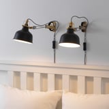 Dvě černé nastavitelné nástěnné lampy RANARP nad bílým dřevěným čelem s polštáři.