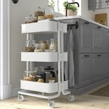 Bílý užitkový vozík IKEA v kuchyni, se sklenicemi a zásobami, vedle skříněk.
