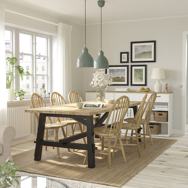 Moderní jídelní kout s dřevěným stolem, židlemi a velkými zelenými závěsnými lampami. Výzdoba zahrnuje zarámované fotografie a vysokou vázu na květiny. Bílé závěsy.