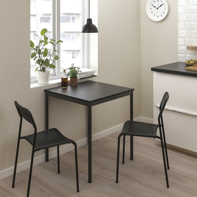 Malý černý sandbergový stůl a židle v rohu u okna. Moderní, prostorově úsporný design s kovovými rámy. Ideální pro malé místnosti.