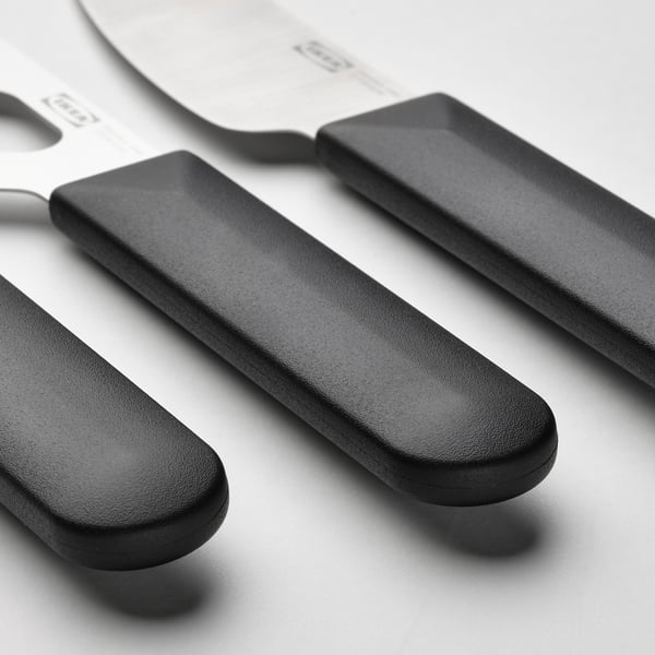 Tři elegantní nože na sýr z nerezové oceli, každý s černou ergonomickou rukojetí, vystavené těsně vedle sebe.