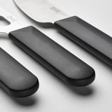 Tři elegantní nože na sýr z nerezové oceli, každý s černou ergonomickou rukojetí, vystavené těsně vedle sebe.