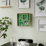 Zelená nástěnná police s nastavitelnými vnitřními policemi, zdobená předměty a rostlinami, obklopená uměním a černou židlí s nádobím.