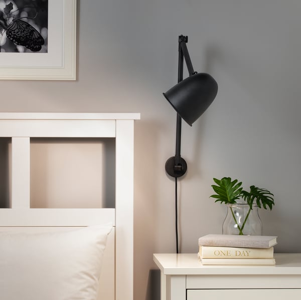 Černá nastavitelná nástěnná lampa nad bílým nočním stolkem s rostlinou a knihou.