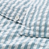 Detail modro-bílého pruhovaného povlečení se zapínáním na knoflík, které předvádí jeho seersuckerovou texturu pro lepší prodyšnost.