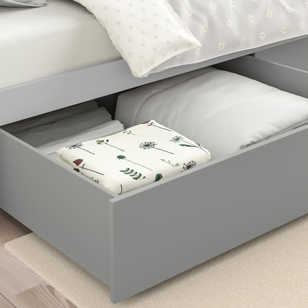 Šedá postel s otevřenou úložnou zásuvkou, která představuje skládané ložní prádlo s květinovým vzorem. Zásuvka se hladce pohybuje na kolečkách.