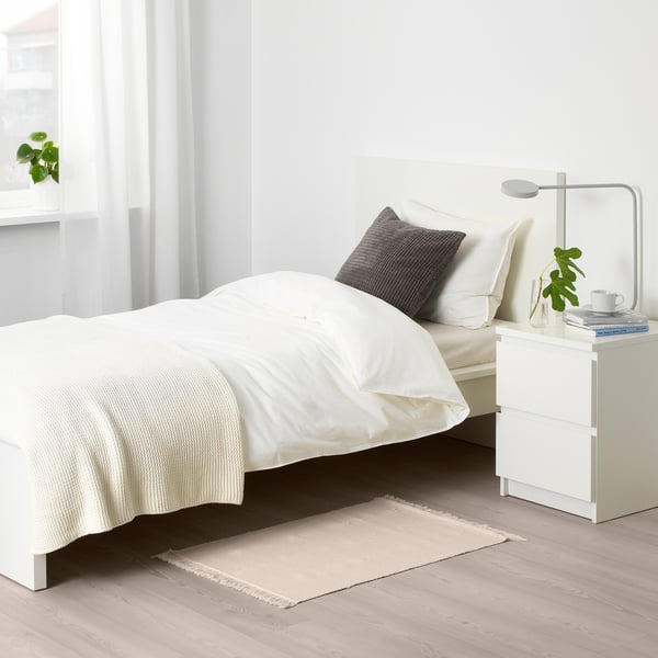 Minimalistická ložnice s béžovým kobercem, bílou postelí a nočním stolkem. Neutrální dekor.
