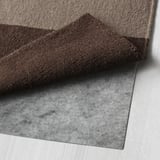 Skládaný hnědý kostkovaný vlněný koberec spočívající na šedé plstěné podložce, předvádějící svou ručně tkanou texturu a vrstvený design.