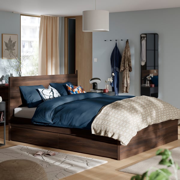 Moderní ložnice – tmavá dřevěná postel STORKLINTA s modrým povlečením, visutými kabáty a velkou závěsnou lampou.