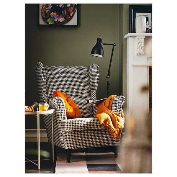 Útulný koutek s křeslem STRANDMON houndstooth, oranžovými polštáři, žlutou dekou, stojací lampou a zelenými stěnami.
