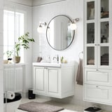 Moderní bílá koupelna s umyvadlem TÄNNFORSEN, velkým kulatým zrcadlem, prosklenými skříňkami a radiátorem. Rostliny a ručníky podtrhují minimalistický design.