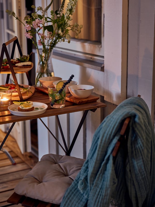 Balkon se stolem TÄRNÖ, jídlo, květiny, osoba v modré dece.