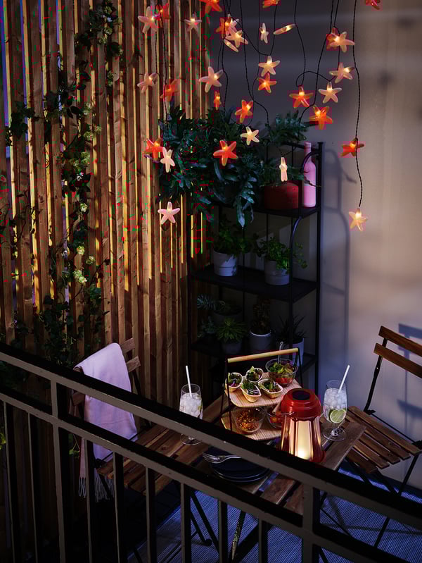 Malý balkon se stolem a židlí TÄRNÖ, zdobený světelnými řetězy, rostlinami a jídlem.
