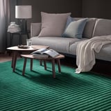 Útulný obývací pokoj s šedou pohovkou, levandulovými/modrými polštáři, odkládacími stolky s vodou a časopisem, tmavě šedými stěnami, zeleným pruhovaným kobercem.