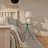 Moderní, malý, tmavě zelený odkládací stolek TÅNEBRO vedle postele, na kterém je lampa a šálek. Stolky kompaktní velikosti jsou ideální do útulných prostor.