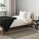 Minimalistická ložnice s černobílým dekorem. Malá dřevěná postel s kobercem TIPHEDE, bílým ložním prádlem, malým stolkem a lampou. Záclony, rostlina, šedá stěna.