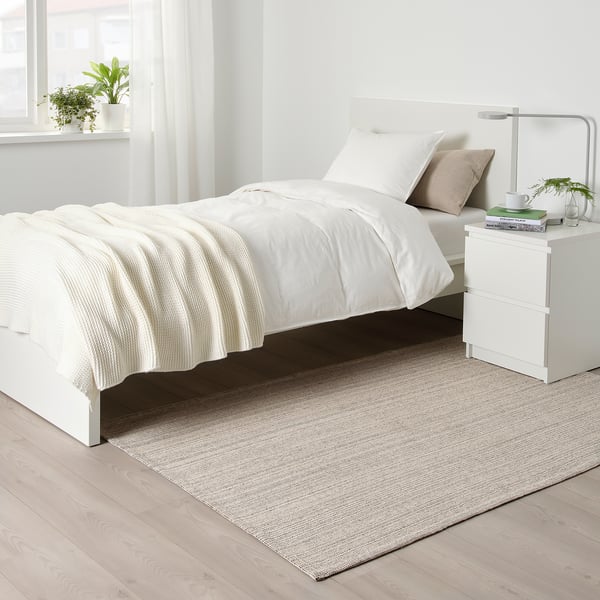 Moderní minimalistická ložnice s kobercem TIPHEDE pod jednolůžkem, bílým povlečením, béžovým polštářem, nočním stolkem s lampou, knihami a rostlinami.