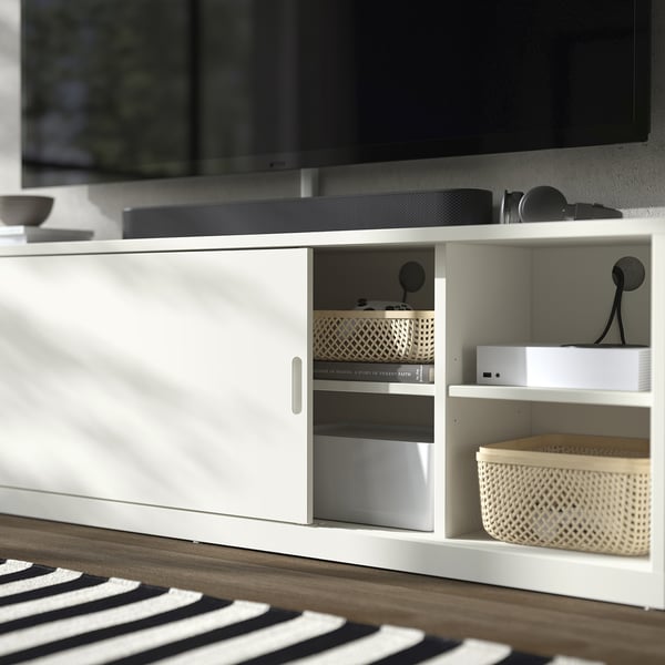 Moderní bílý tv stolek s posuvnými dveřmi, košíkem a elektronikou. Čistý, organizovaný design.