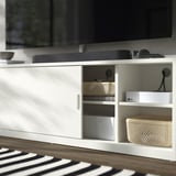 Moderní bílý tv stolek s posuvnými dveřmi, košíkem a elektronikou. Čistý, organizovaný design.