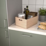Světlá dřevěná krabička na pultu mezi zelenými skříňkami s lahvemi, plechem, dřevěnými deskami, bylinkami.