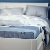 Detailní záběr na modrou matraci IKEA na bílé posteli. Viditelná je cedulka na matraci s modrobílým designem.