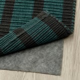 Tmavě zelený a černý pruhovaný koberec, umístěný na šedé podložce na dřevěné podlaze. Koberec má rovný povrch, takže vysávání je snadné.