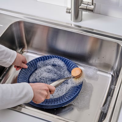 Osoba myjící modré nádobí ve dřezu pomocí štětce na nádobí a mýdla.