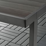 Detail rohu moderních venkovních stolů s deskou připomínající tmavé dřevo a kovovým rámem s práškovým nástřikem.