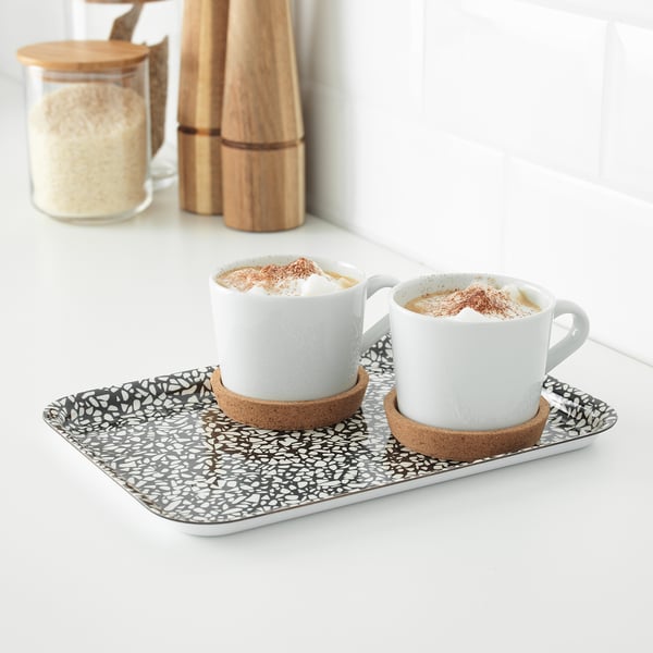 Dva hrnky cappuccina na vzorovaném podnosu s korkovými tácky, položené na bílé desce.