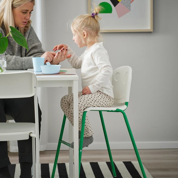 Mladá dívka sedí na zelené a bílé židli a jí z modré misky u bílého stolu se ženou opodál.
