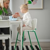 Mladá dívka sedí na zelené a bílé židli a jí z modré misky u bílého stolu se ženou opodál.