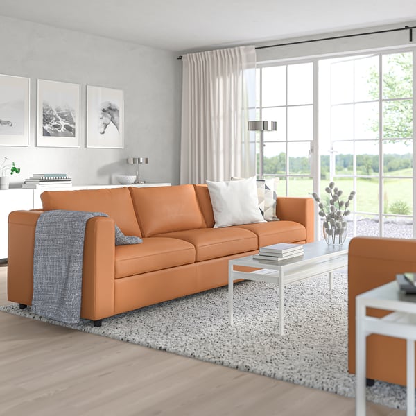 Moderní obývací pokoj s oranžovou pohovkou VIMLE, mix kůže a látky. Přirozené světlo naplňuje minimalistický prostor bílým dekorem.