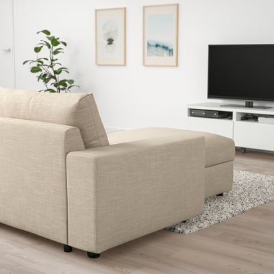 Pohovka VIMLE ve světle béžové barvě s lenoškou směřující doleva, TV, rostlinou, minimalistickým dekorem.