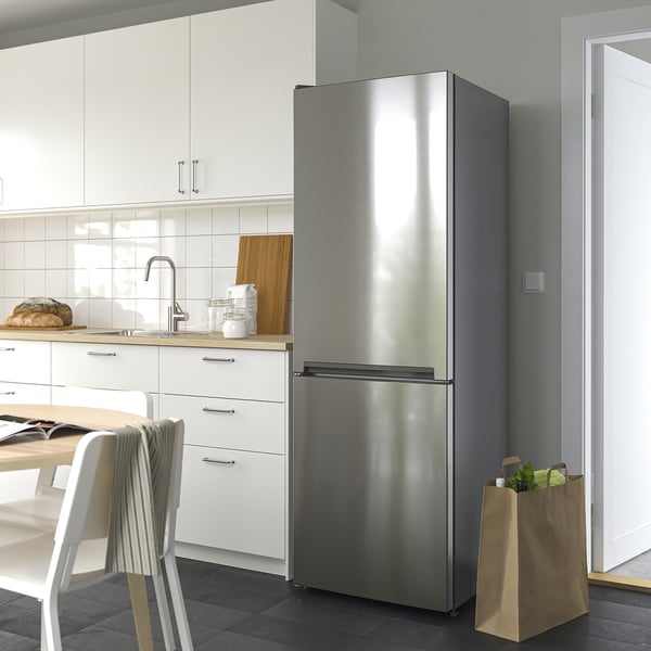 Moderní kuchyňská linka s nerezovou dvoudveřovou lednicí VINDÅS, která je větší než židle a skříňky v okolí, má elegantní design a skleněné police.