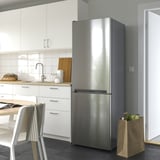 Moderní kuchyňská linka s nerezovou dvoudveřovou lednicí VINDÅS, která je větší než židle a skříňky v okolí, má elegantní design a skleněné police.