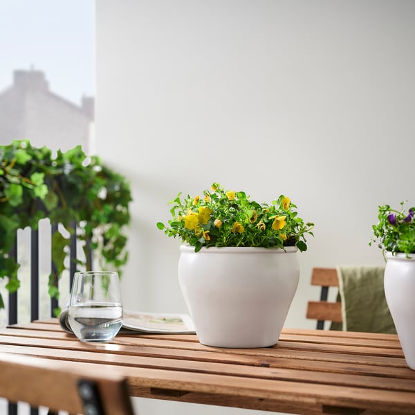 Balkonová scéna s bílými květináči se žlutými květy na dřevěném stole, doplněnými sklenicemi na víno a zelenou rostlinou.
