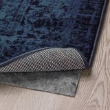 Roh tmavě modrého vzorovaného koberce s texturovanou spodní stranou pro uchopení je zobrazen na dřevěné podlaze.