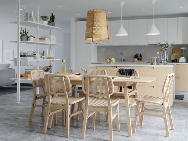 Světlá, minimalistická kuchyň se světlou dřevěnou jídelní soupravou s tkanými židlemi, závěsnými proutěnými lampami a bílými závěsnými svítidly.