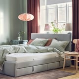 Cozy bedroom with grey ÅBYGDA mattress, light-colored pillows, pink lampshade, wooden nightstands, and large window with red curtains.