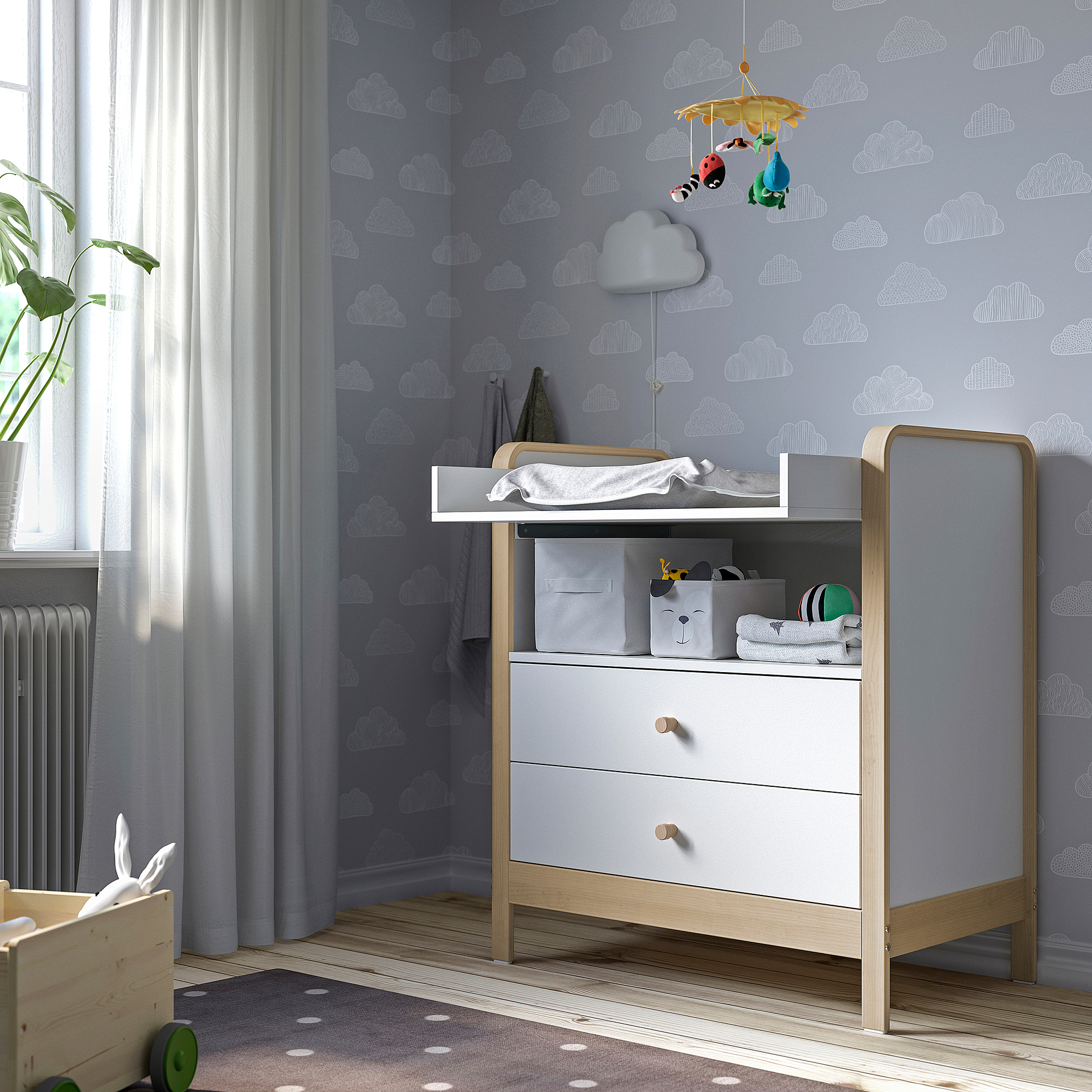 Grey nursery dresser with 3 drawers, changing top, mobile, plant, blue wallpaper, rug.