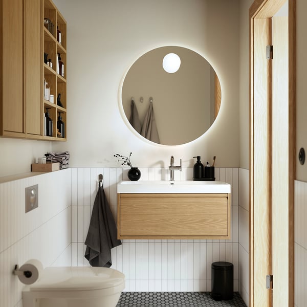Modern bathroom: round mirror, oak sink, marble top, black accessories, white toilet, grey towels.
