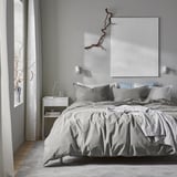 Modern bedroom with grey ÄNGSLILJA duvet on bed, soft recycled cotton. Large white art above, plant on small white bedside table, grey wall.