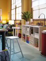 Bright room with a yellow wall, a white junior chair, toy shelf, and a wooden table. Large windows let in natural light.