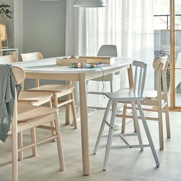 Modern dining table with light wood chairs. Craft tools and paper on table. Large window with sheer curtains. Minimalist decor, airy room.