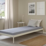 Gray foam mattress on white bed frame in minimalist room with plant and lamp.