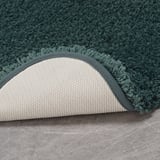 Green bath mat with fluffy texture and cream-colored non-slip backing, placed on a gray floor.