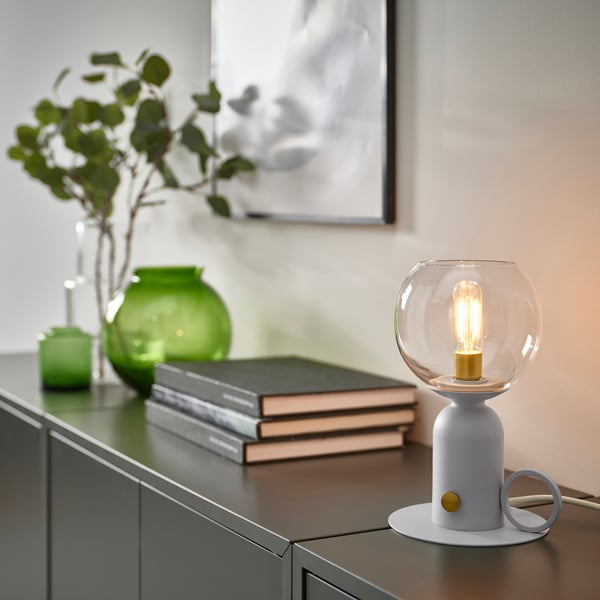 A modern gray ÅSKMULLER table lamp with a clear glass shade, sitting on a shelf next to a stack of books and green vases, casting a warm glow.